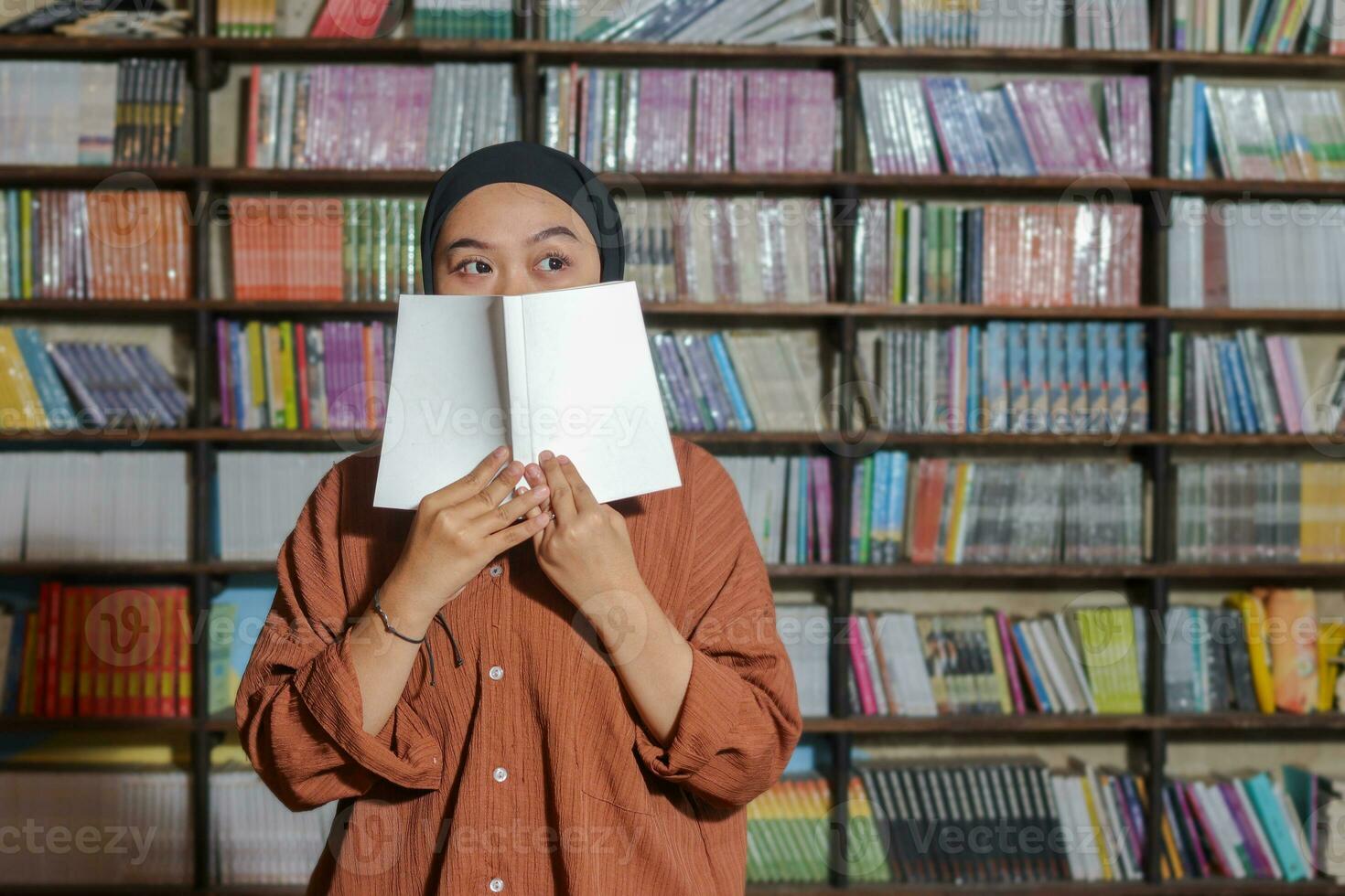 retrato do ásia hijab mulher cobertura dela metade cabeça com uma livro dentro frente do biblioteca estante. muçulmano menina lendo uma livro. conceito do alfabetização e conhecimento foto