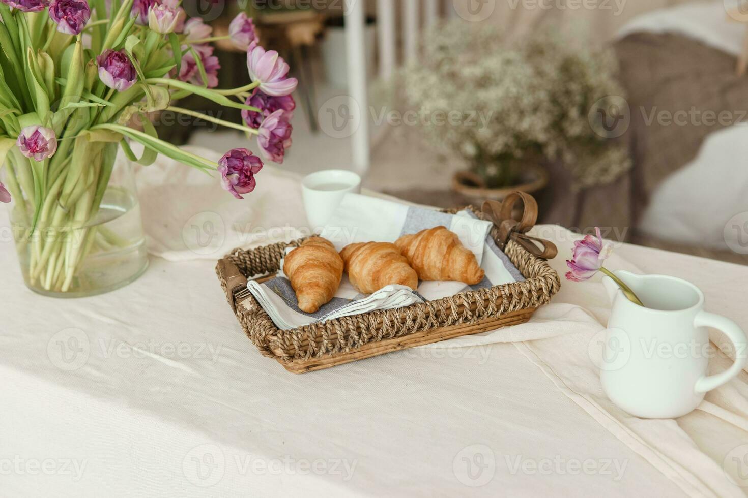 fresco croissants dentro uma vime cesta em a mesa Próximo para uma vaso do lilás tulipas. foto