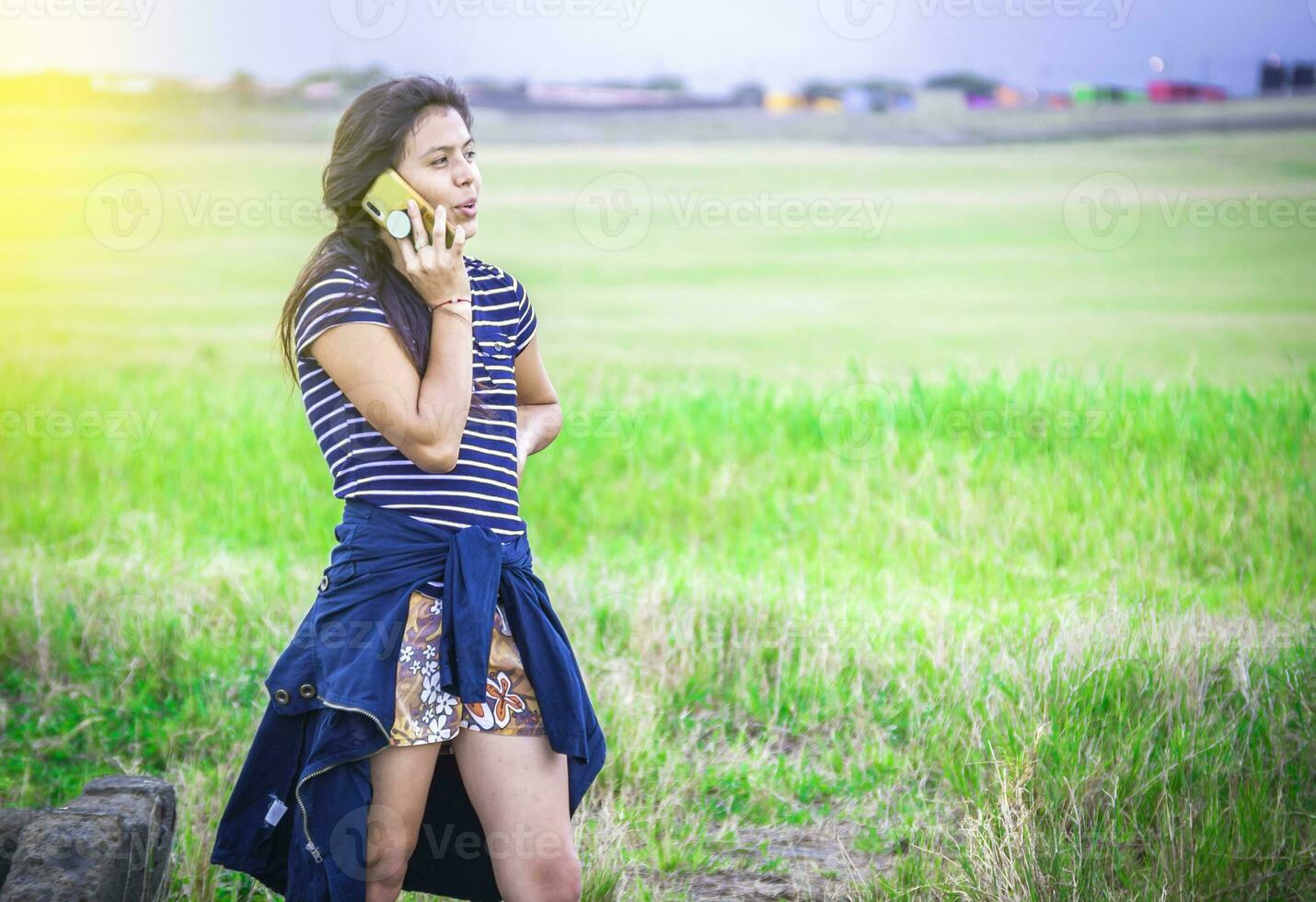 jovem menina chamando telefone em a verde campo, lindo jovem mulher chamando de célula telefone em a Relva ar livre. foto