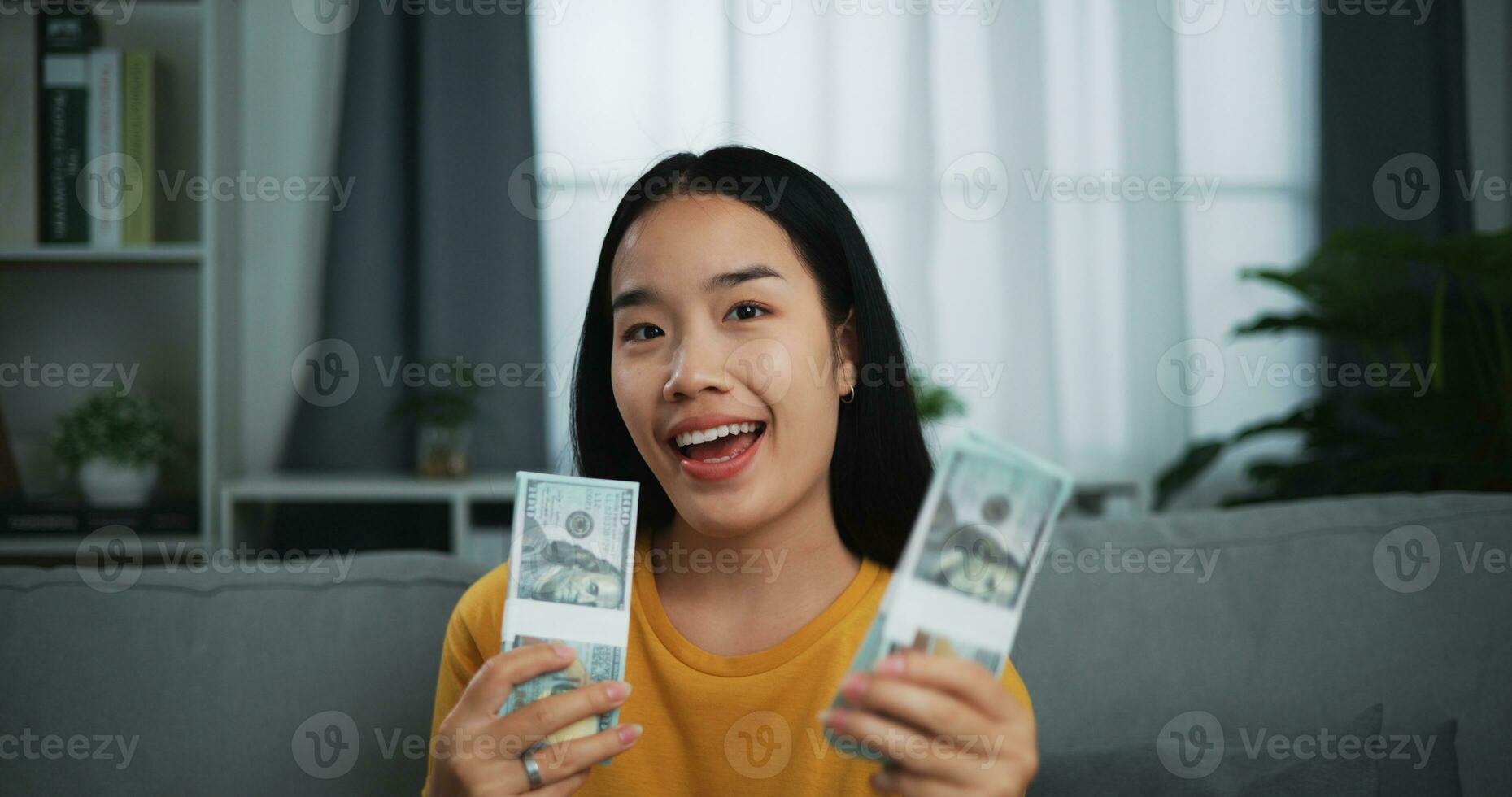 retrato do jovem ásia mulher mostrando dólar às Câmera e sorridente em sofá dentro a vivo quarto às lar. foto