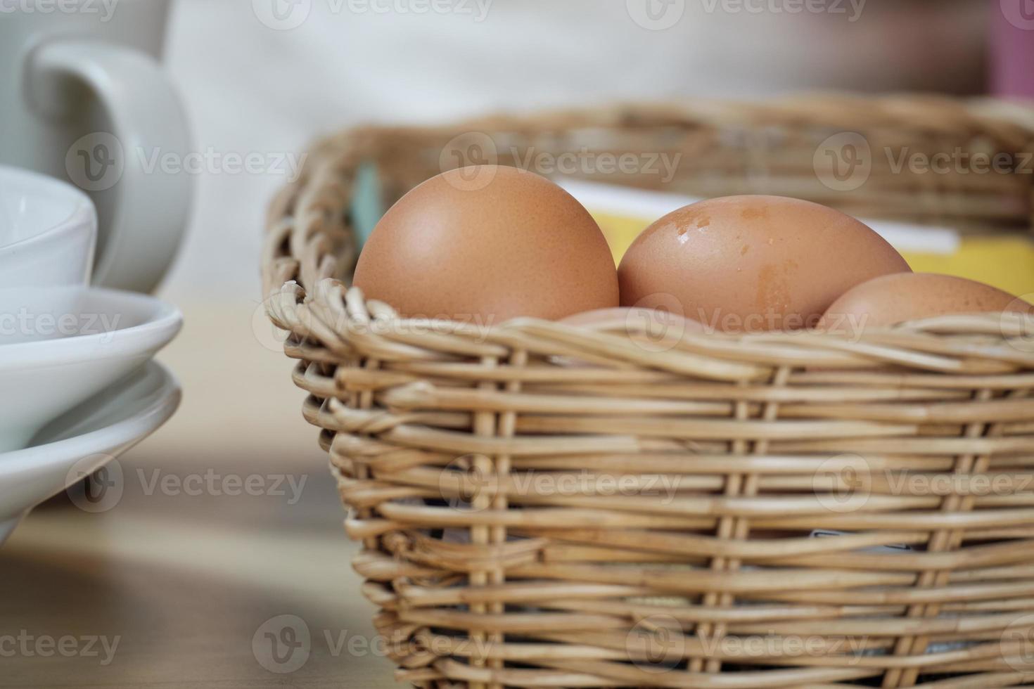ovos frescos orgânicos na cesta tecida, copos brancos sobre uma mesa de madeira. foto