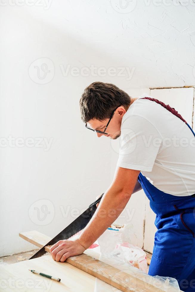 carpinteiro de macacão azul serrando uma tábua com uma serra manual foto