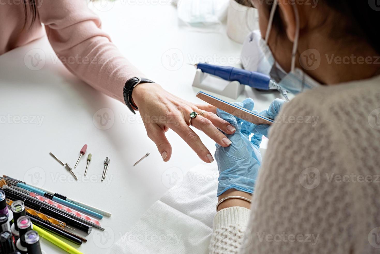 Mestre de manicure em luvas usando lixa de unha para polir a unha de uma cliente foto