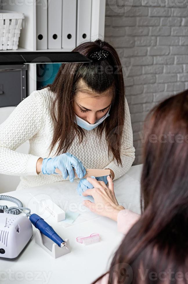 Mestre de manicure usando lixa de unha para polir a unha de uma cliente foto