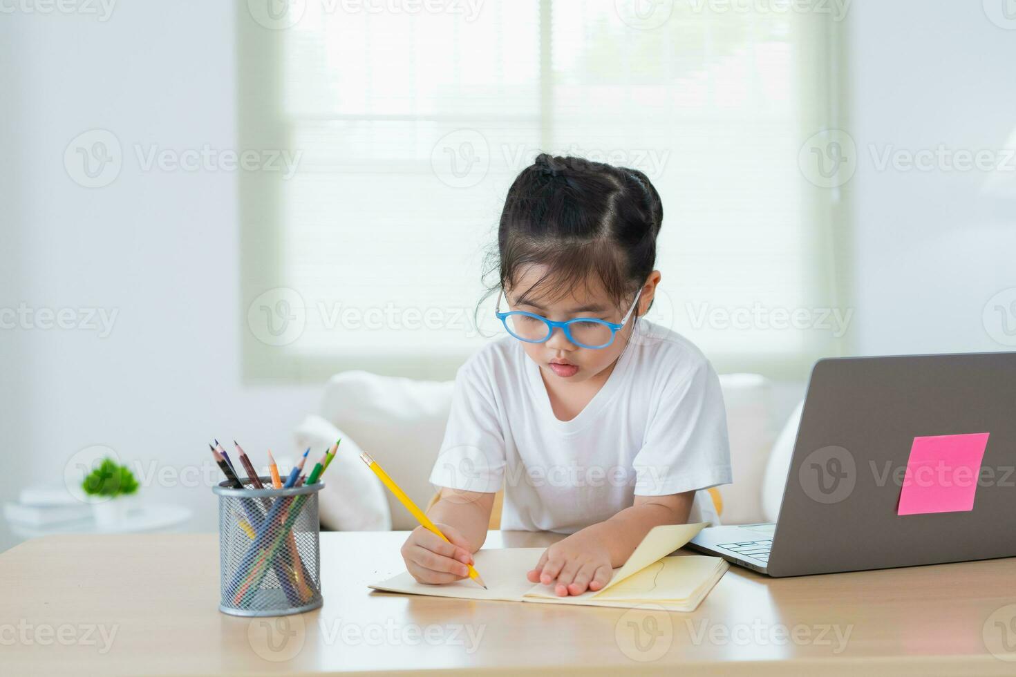 ásia bebê menina vestindo Óculos fazendo casa usando computador portátil e escrever notas dentro caderno para estude conectados em madeira mesa escrivaninha dentro vivo quarto às lar. Educação Aprendendo conectados a partir de casa conceito. foto