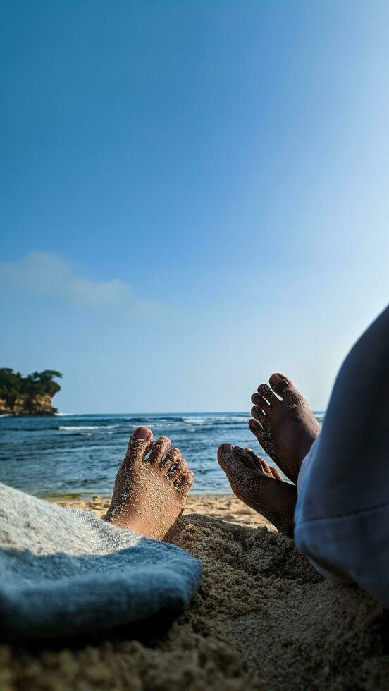 a casal é desfrutando seus período de férias às a praia, deitado em a areia e olhando Fora às a oceano. a do casal pés estão coberto dentro de praia areia. verão e de praia conceito. foto