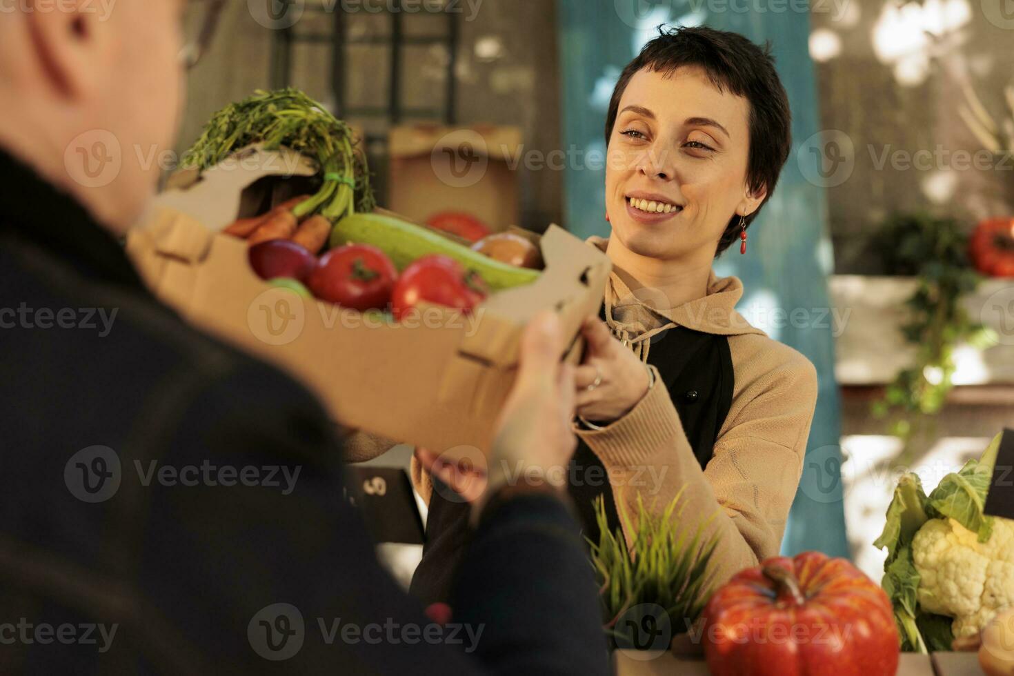 jovem agradável mulher dando caixa cheio do fresco orgânico frutas e legumes para cliente enquanto trabalhando às local agricultores mercado. fêmea agricultor vendendo saudável localmente crescido Fazenda Comida foto