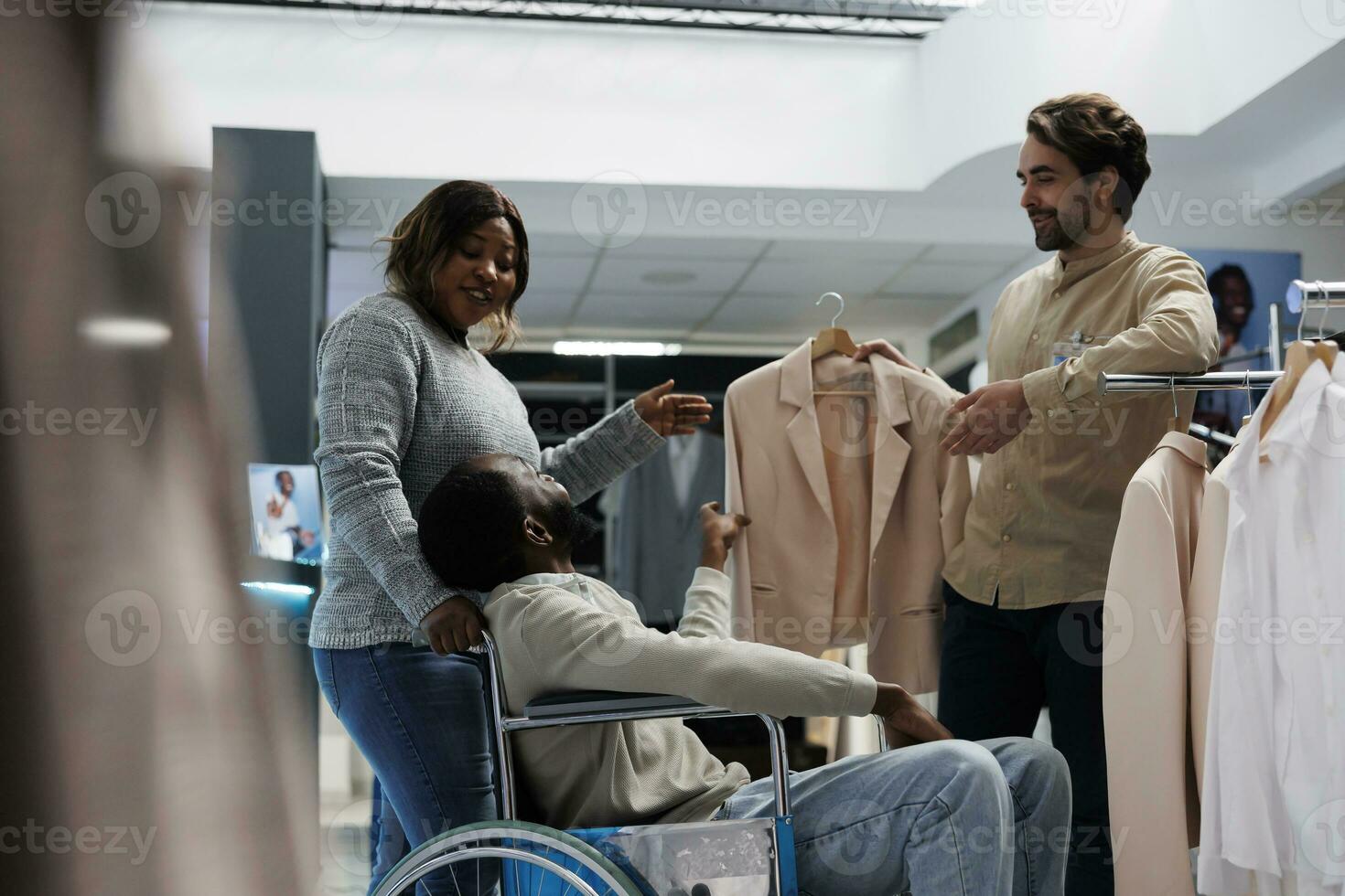 homem com físico incapacidade escolhendo Jaqueta com namorada enquanto compras dentro roupas loja. departamento Shopping africano americano cliente dentro cadeira de rodas selecionando equipamento com esposa foto