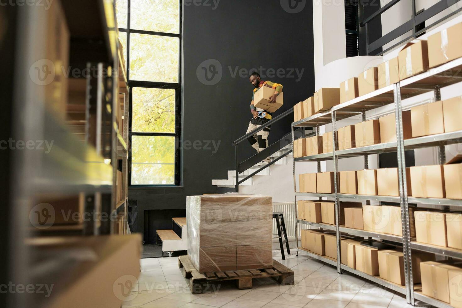 africano americano trabalhador carregando cartão caixa em escadas durante armazém inventário, preparando clientes ordens. armazenamento quarto trabalhando empregado com mercadoria produtos dentro armazém. varejo conceito foto