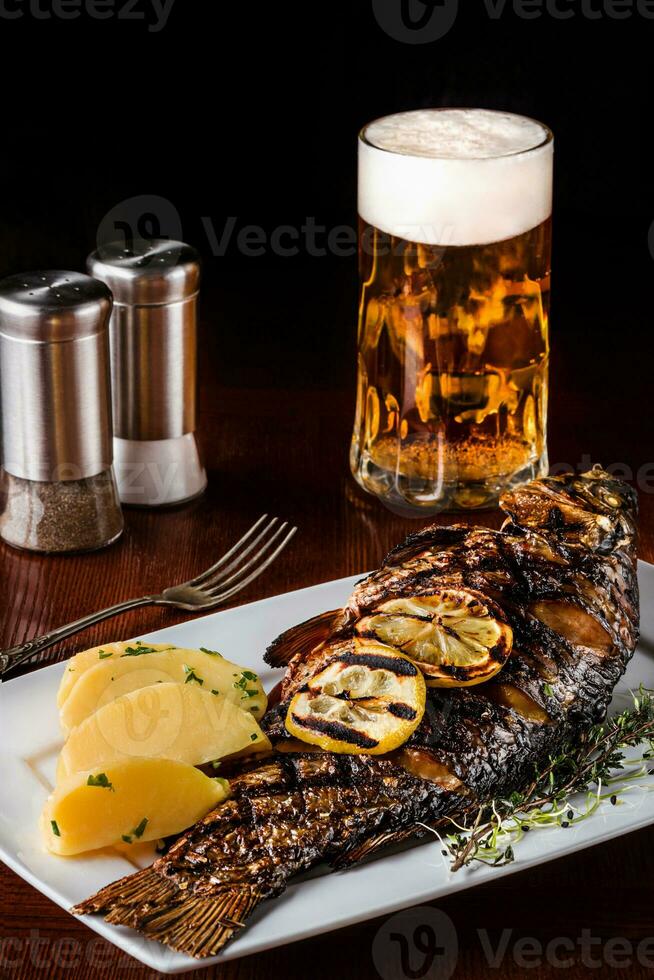 grelhado carpa peixe com alecrim batatas, limão e Cerveja em uma de madeira mesa. Preto fundo. foto