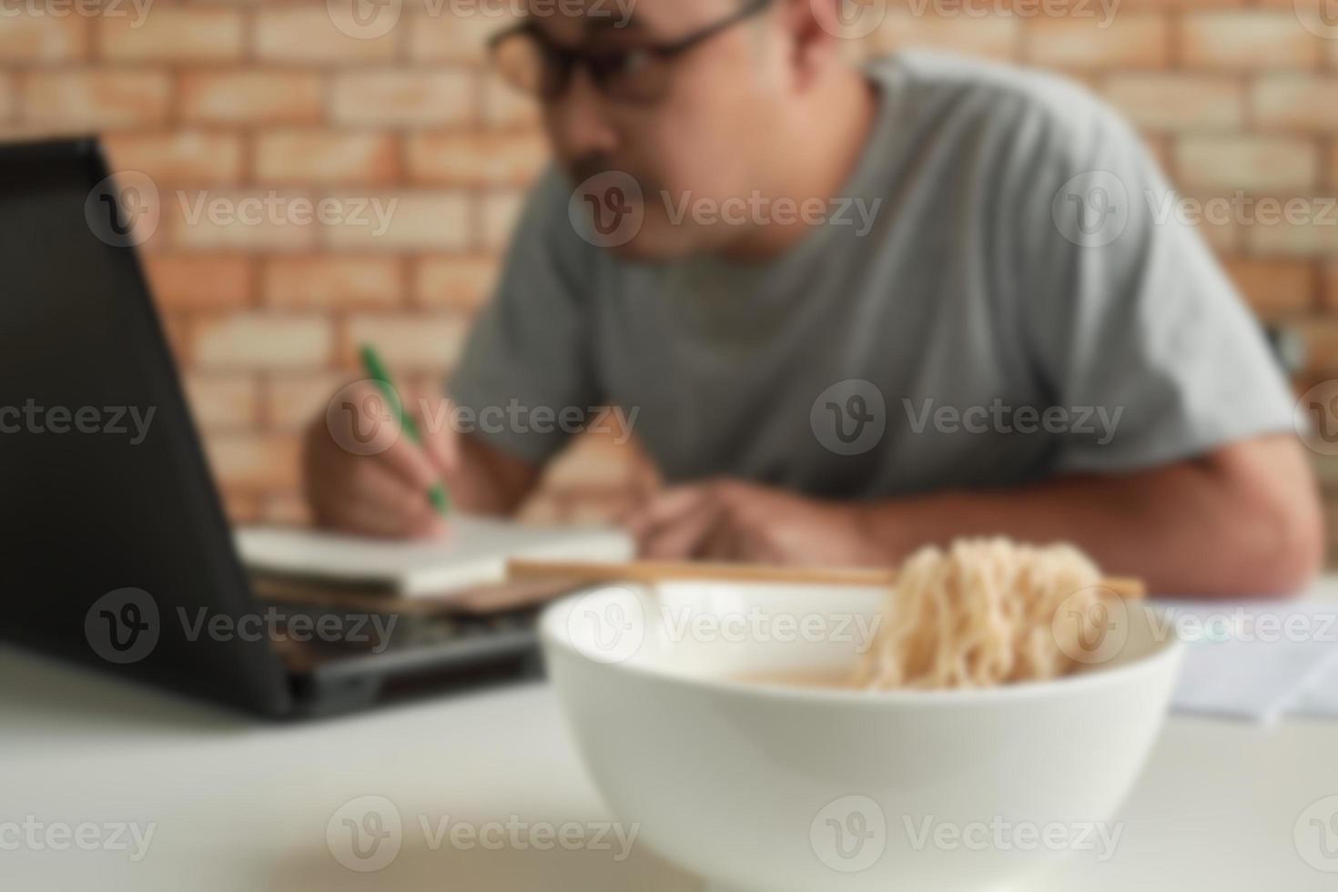 homem trabalha com um laptop e come macarrão instantâneo durante as pausas para o almoço. foto