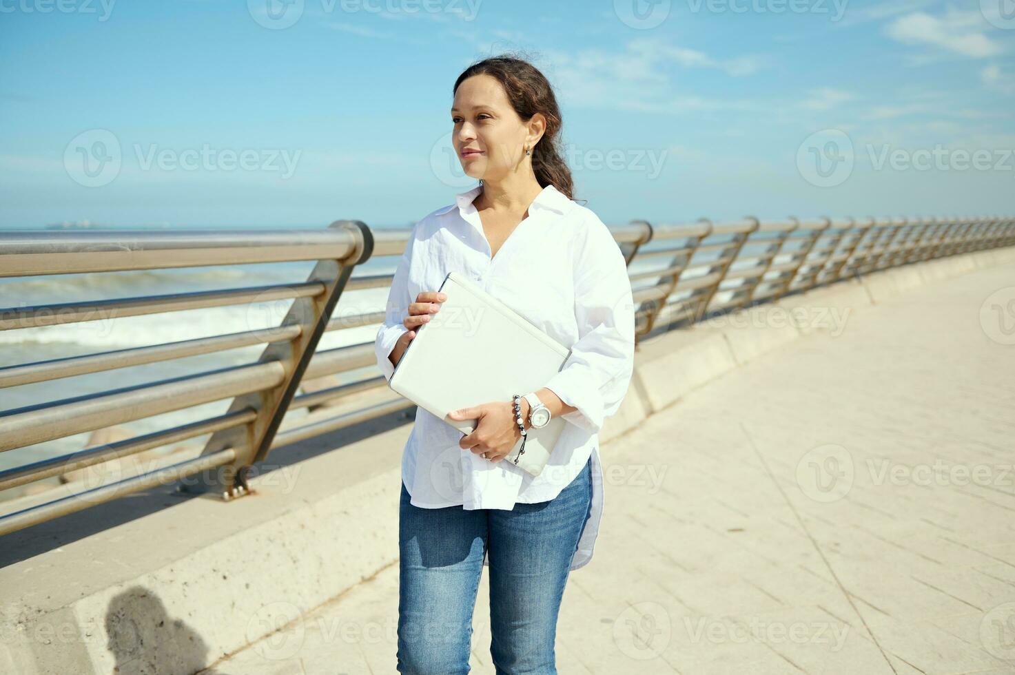 confiante jovem mulher caminhando ao longo a passeio, sonhadoramente olhando aparte, segurando computador portátil, planejamento Novo comece projetos foto