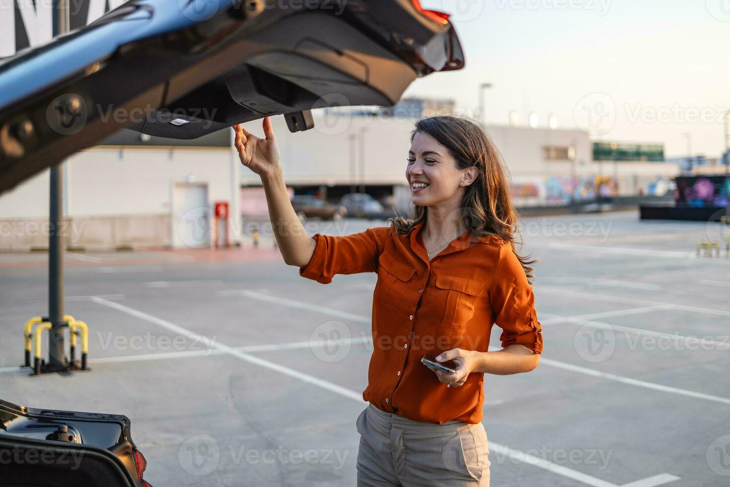 jovem lindo elegante mulher motorista é abertura carro tronco sorridente às estacionamento muito, mulher fechamento carro tronco estacionado em pé de a costas tronco do dela carro em a estacionamento muitos foto