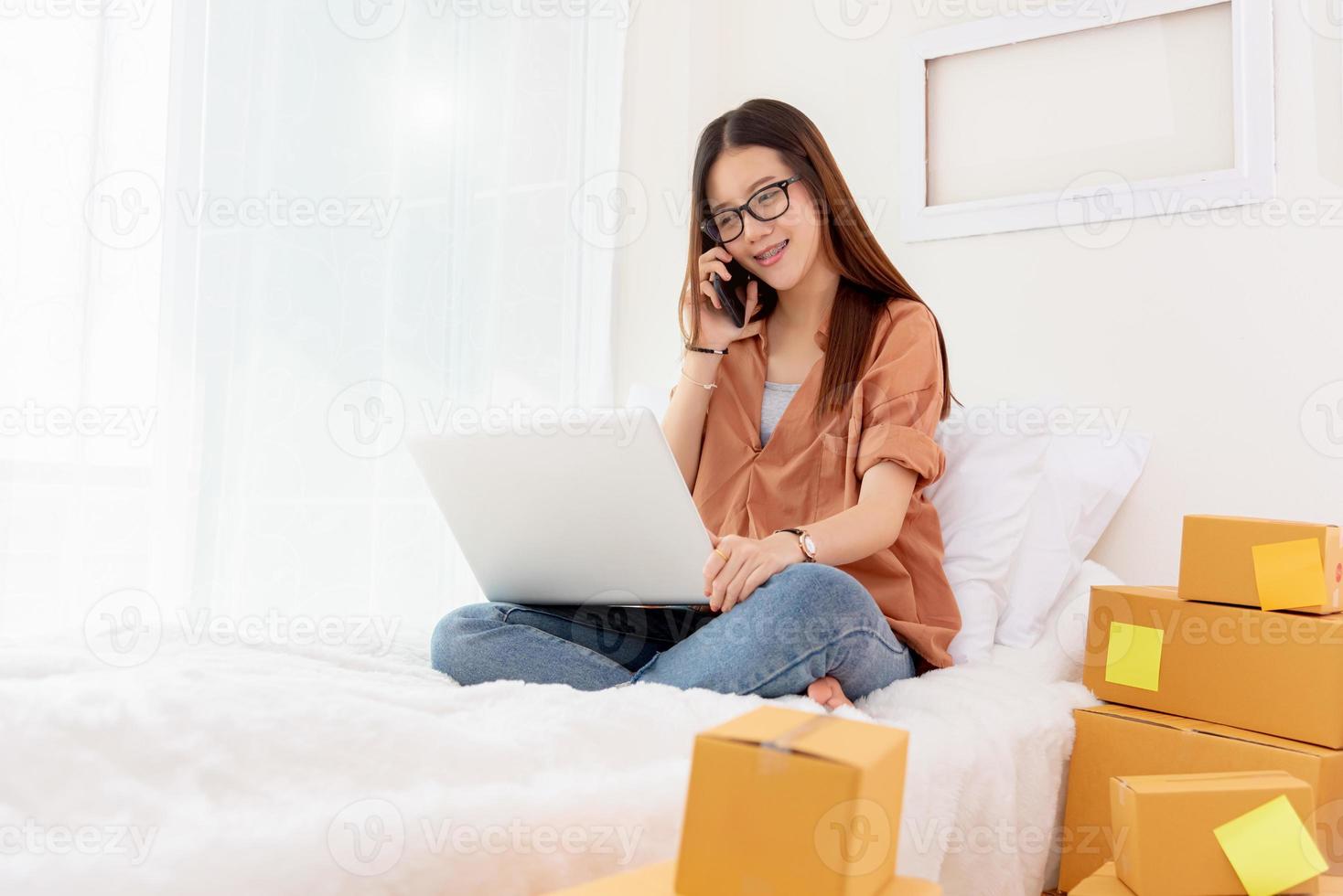 Mulher asiática bonita usando laptop e ligando para o telefone na cama foto