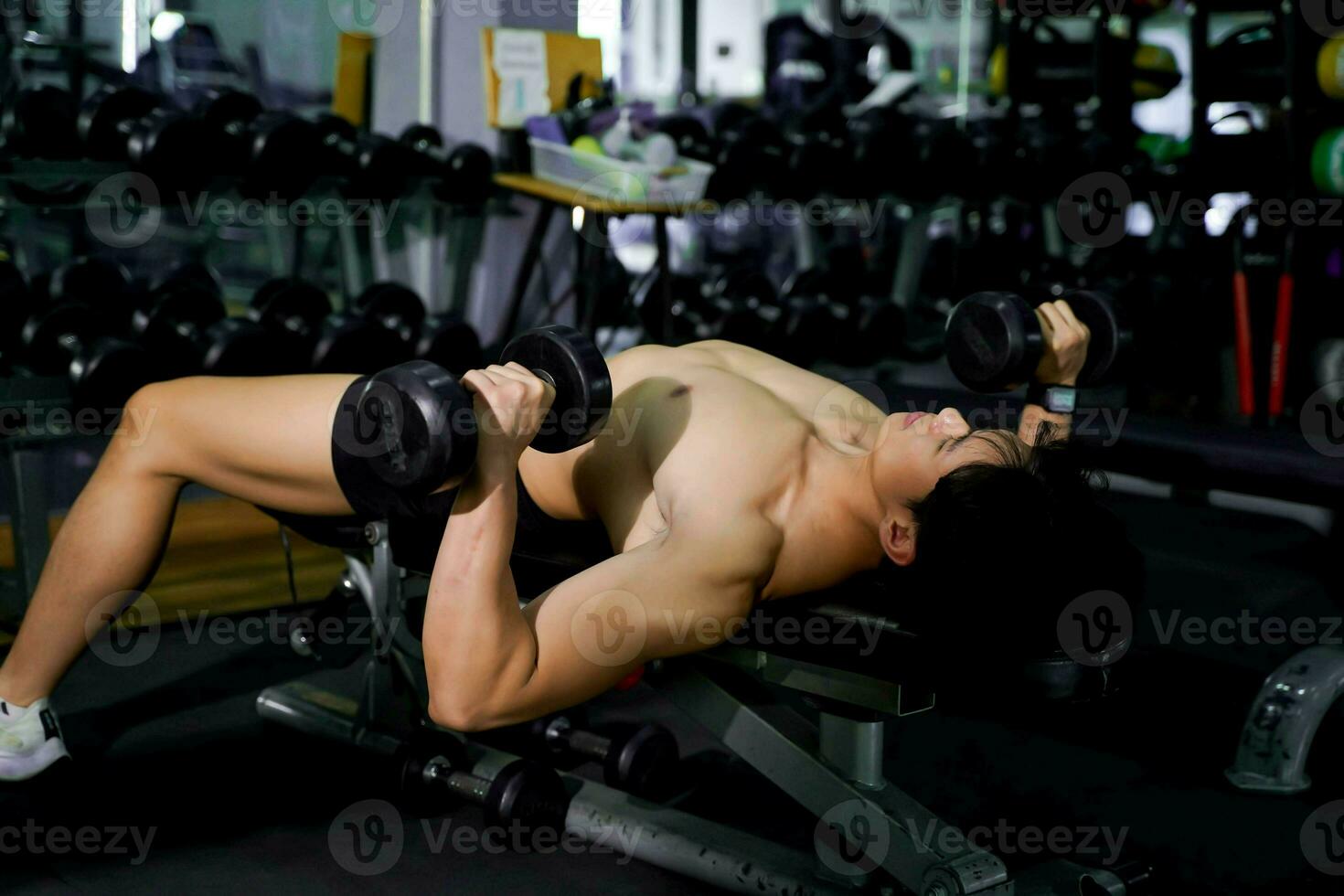 fechar-se ásia bonito homem com perfeito corpo jogando dumbell peso Treinamento dentro a deitado posição para Construir ombro e peito músculos às ginástica Centro. foto