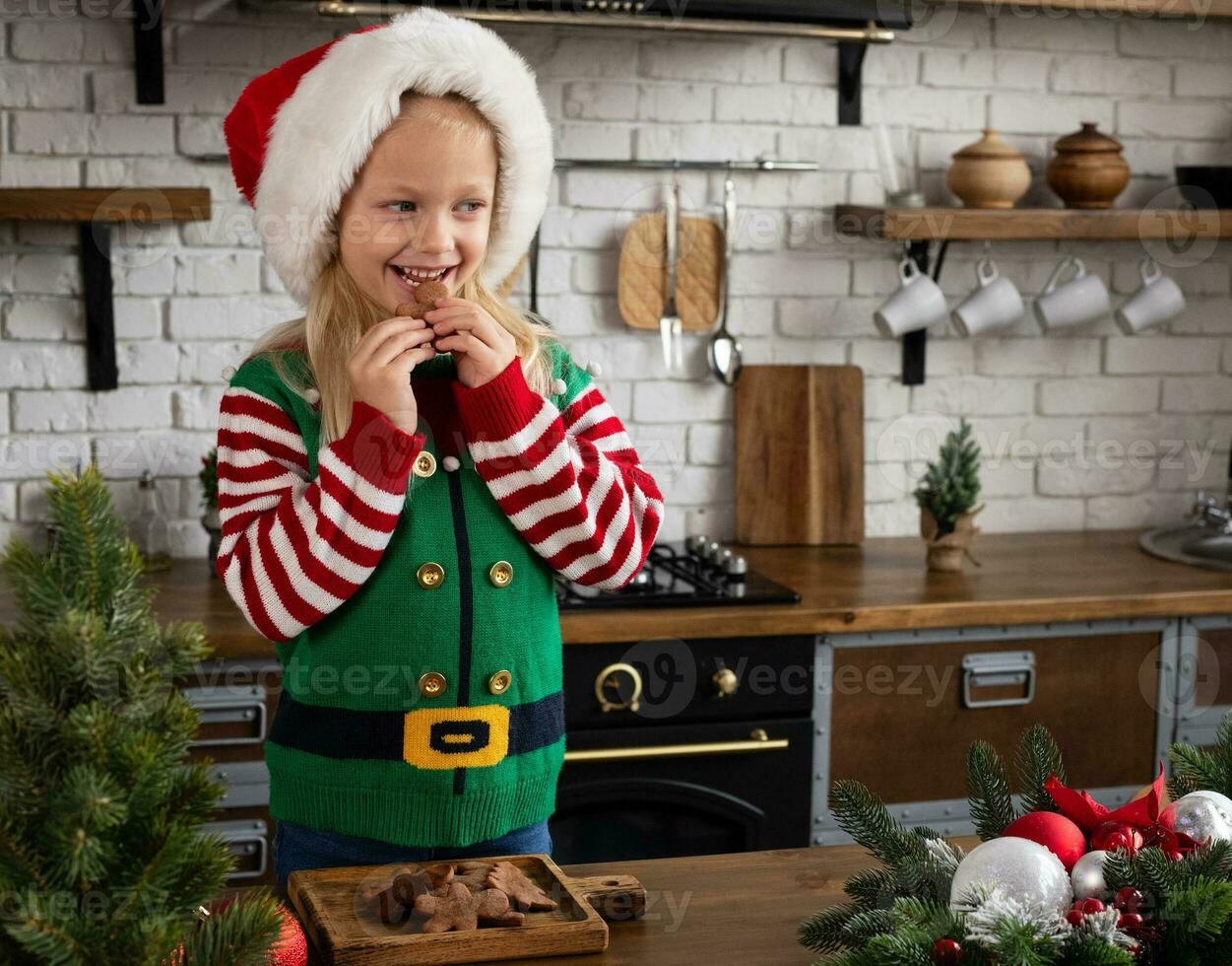 criança dentro santa chapéu, vestido acima Como duende, comendo Pão de gengibre homem bolacha às Natal decorado cozinha. criança olhando a parte, de lado foto