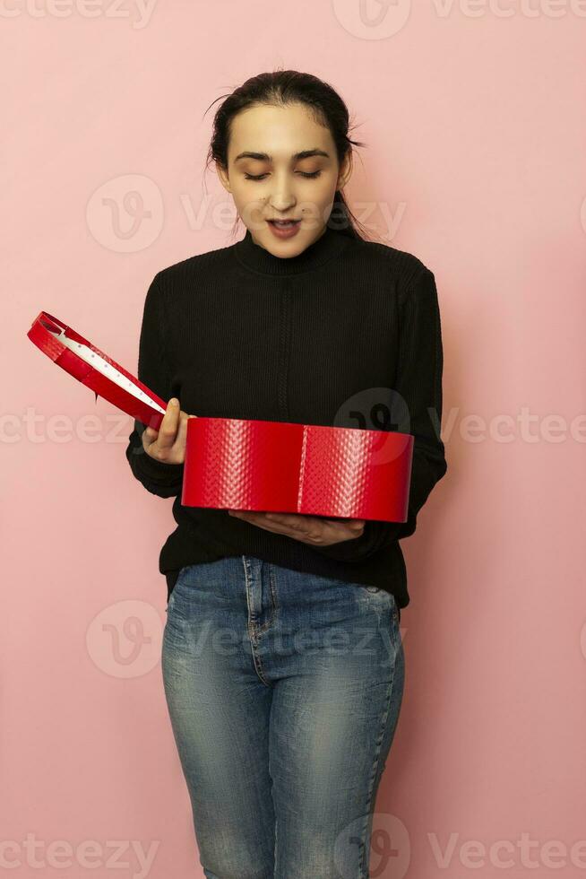 facial emoções. espantado, surpreso mulher detém coração forma presente caixa. atraente moda modelo dentro casual roupas posando com vermelho presente caixa. dia dos namorados dia presente. amor dia, aniversário foto