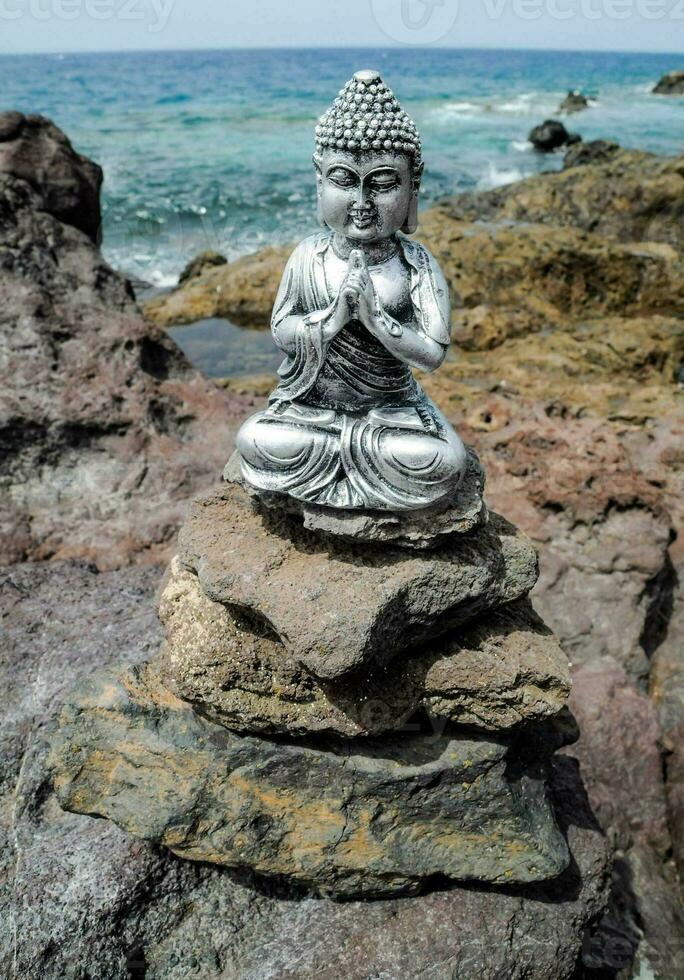 uma prata Buda estátua sentado em topo do pedras de a oceano foto