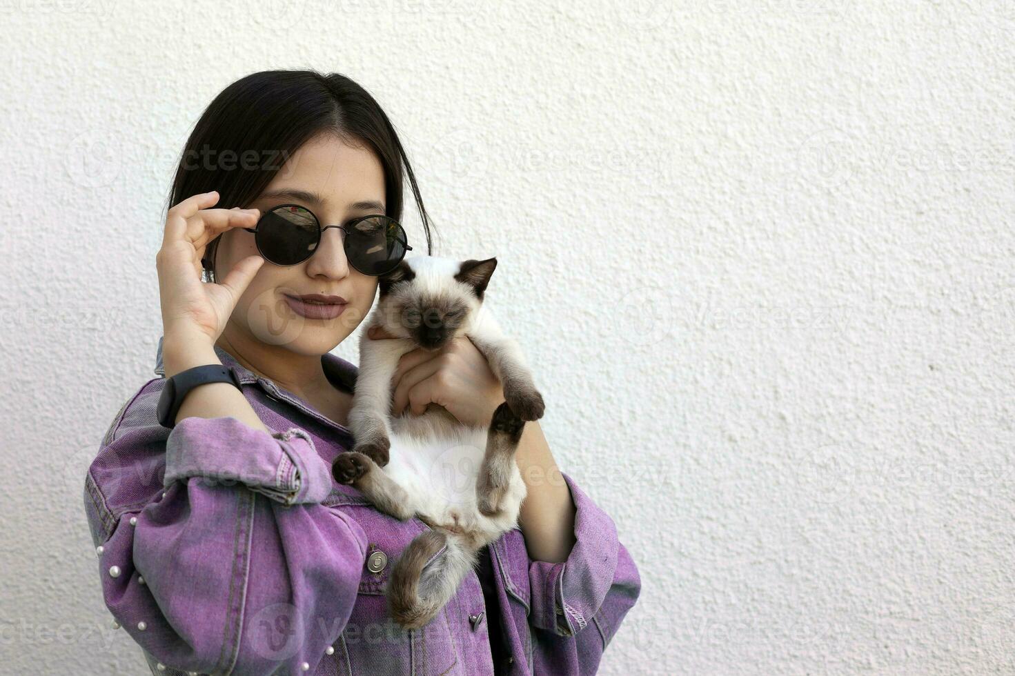 jovem atraente mulher abraçando bichano gato dentro mãos. fofa e glamouroso menina dentro na moda oculos de sol posando com dela siamês gato foto