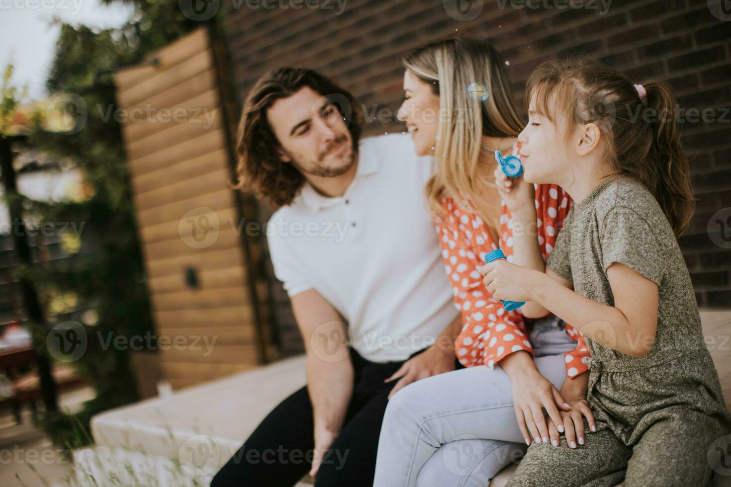 família com uma mãe, pai e filha sentado lado de fora em a passos do uma frente varanda do uma tijolo casa foto