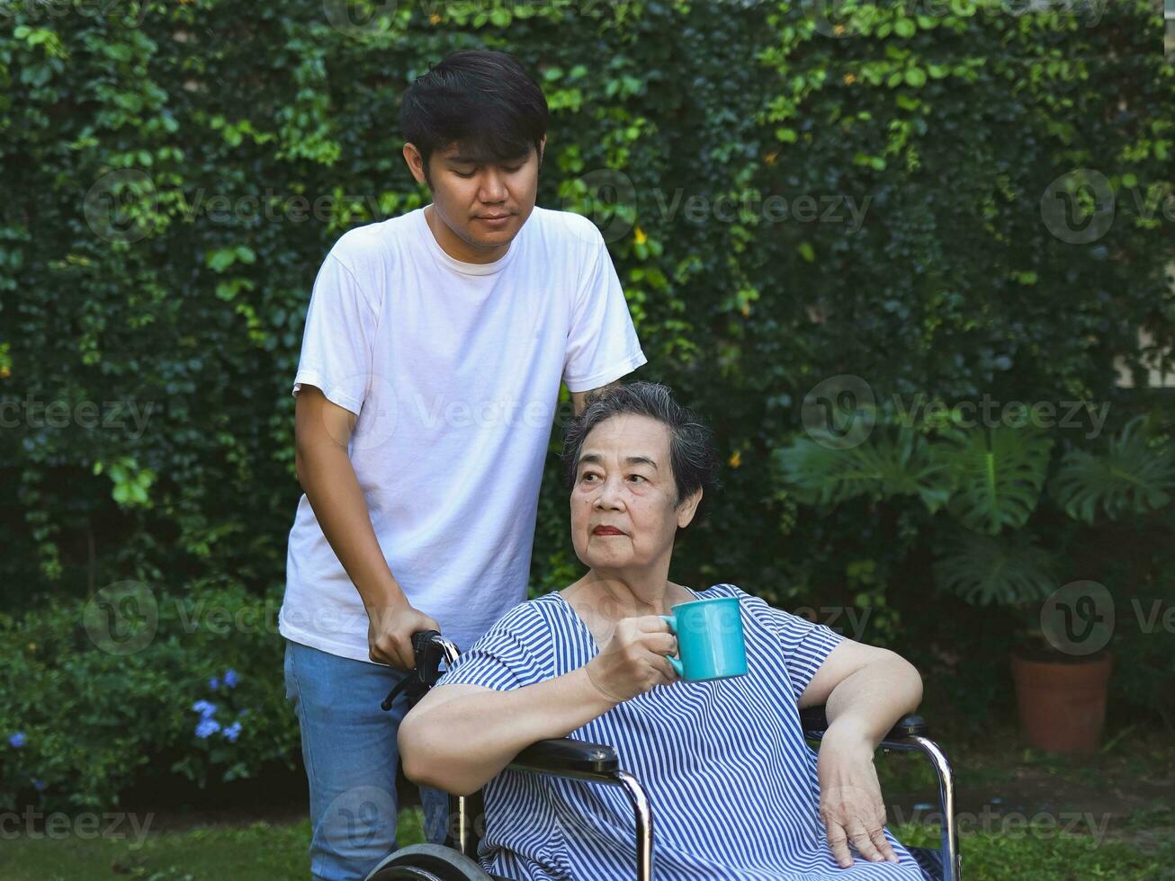 retrato do ásia Senior mulher sentado em cadeira de rodas, segurando azul copo do chá ou café dentro a jardim, com levando Cuidado do dela filho. foto