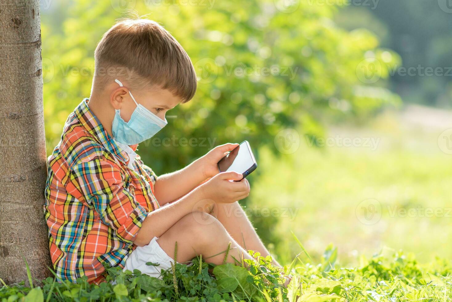 uma criança dentro uma médico mascarar senta em a Relva e parece dentro a telefone desenhos animados dentro a verão às pôr do sol. criança com uma Móvel telefone dentro dele mãos. prevenção contra coronavírus covid-19 durante uma pandemia foto