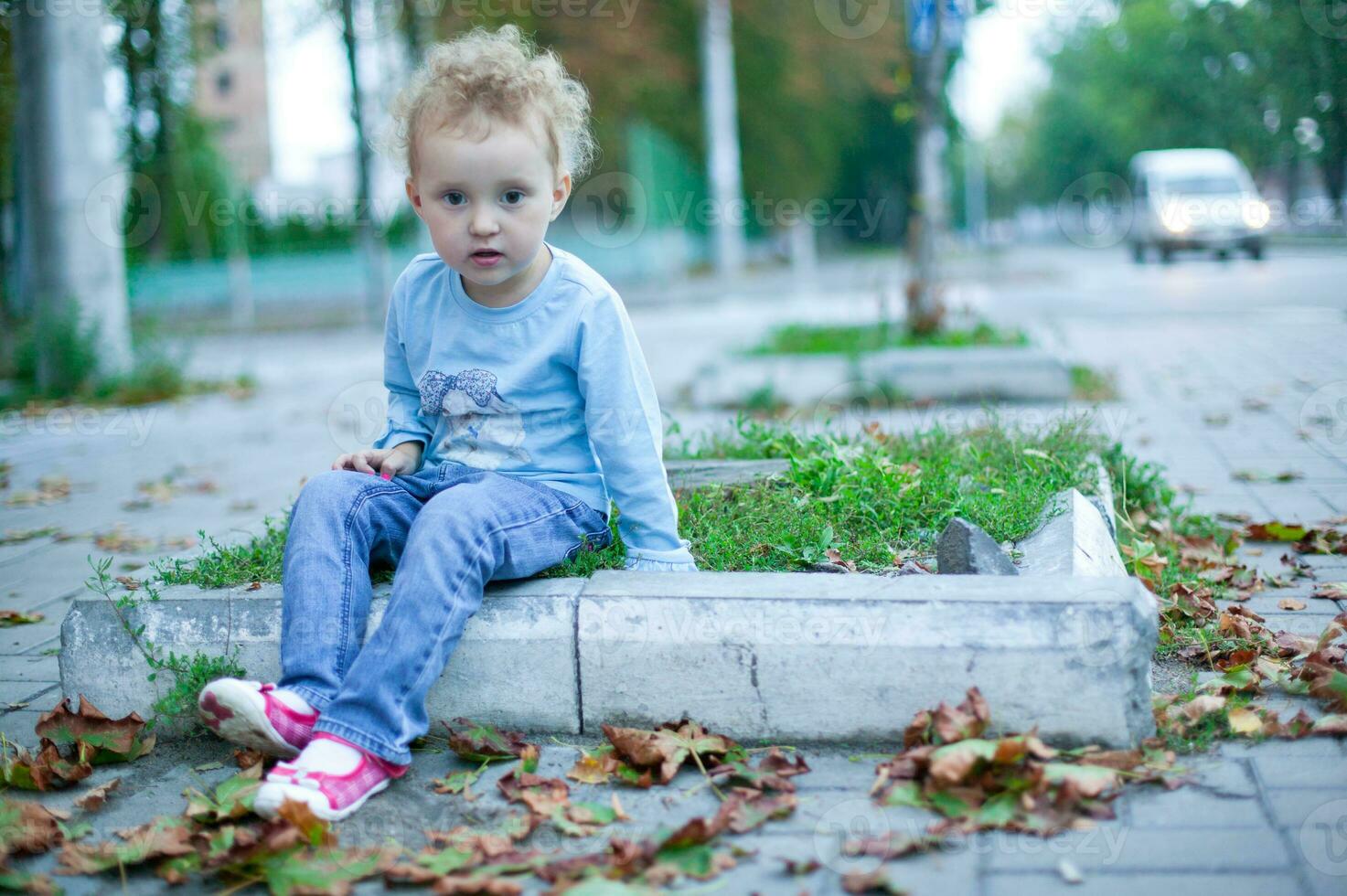 assustado lindo menina com encaracolado cabelo sentado em a borda da calçada dentro a estrada. criança dentro jeans e uma azul camisa jogando ao ar livre foto