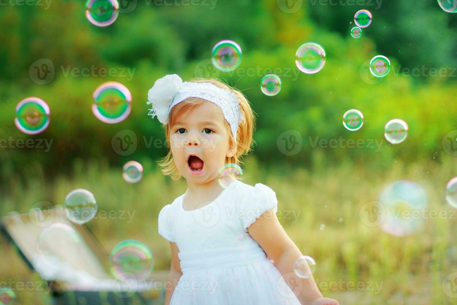 uma pequeno menina dentro uma branco vestir e com uma flor em dela cabeça é feliz com Sabonete bolhas. a criança pega Sabonete bolhas. bebê caminhando dentro a parque foto