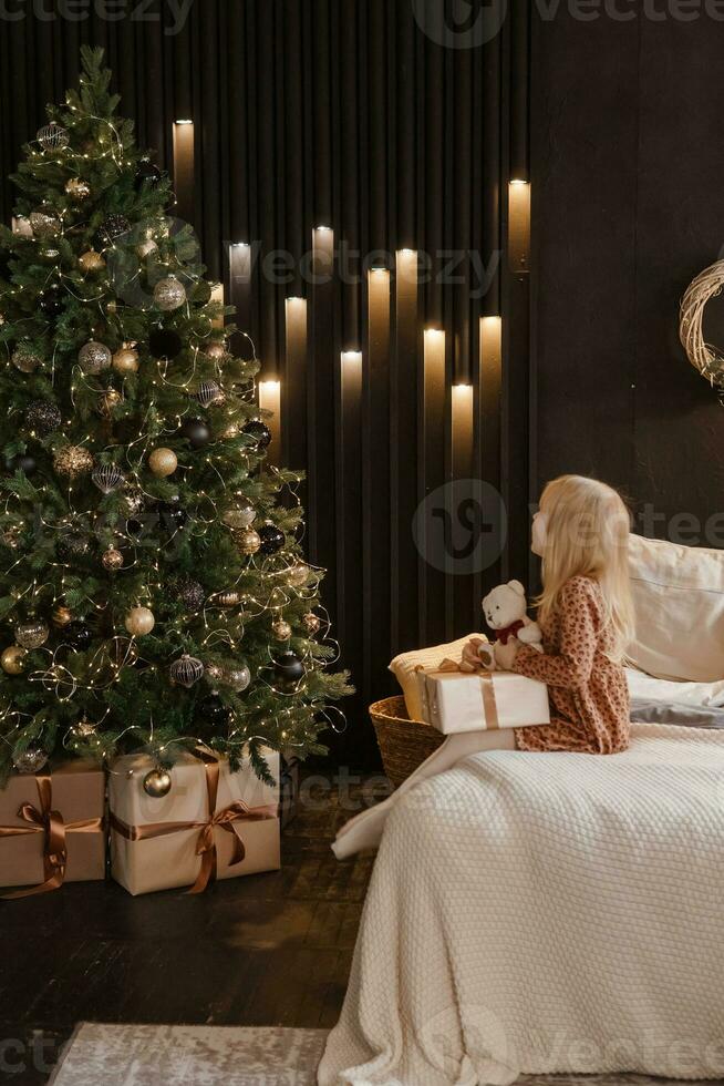 uma pequeno Loiras menina é sentado em uma suave cama dentro uma festivo interior decorado dentro uma Novo anos estilo. uma criança detém uma Natal presente dentro uma construir pacote. foto