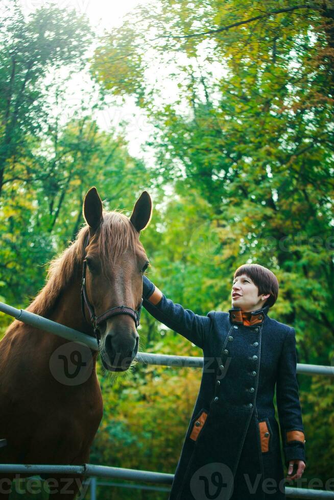 uma mulher dentro uma azul grandes casaco e com curto Sombrio Castanho cabelo estendido dela mão para acidente vascular encefálico a cavalo. a cavalo picado acima dele ouvidos. uma trem para cavalos contra a fundo do uma verde floresta. foto