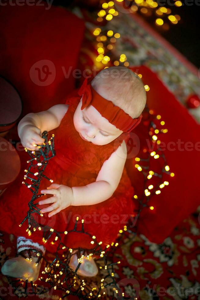 pequeno menina dentro vermelho vestir contra fundo do Natal árvore detém Natal festão dentro dela mãos. bebê 6 mês velho comemora Natal. foto