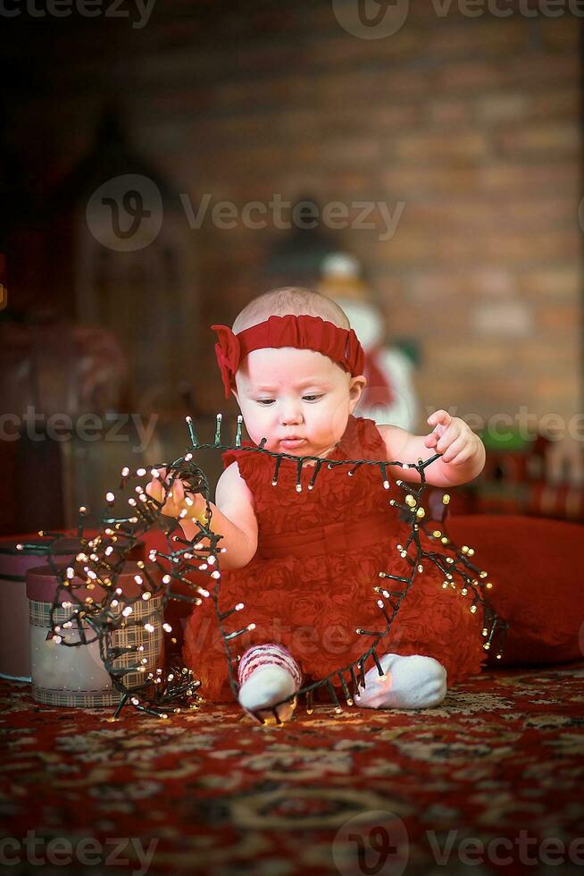 pequeno menina dentro vermelho vestir contra fundo do Natal árvore detém Natal festão dentro dela mãos. bebê 6 mês velho comemora Natal. foto