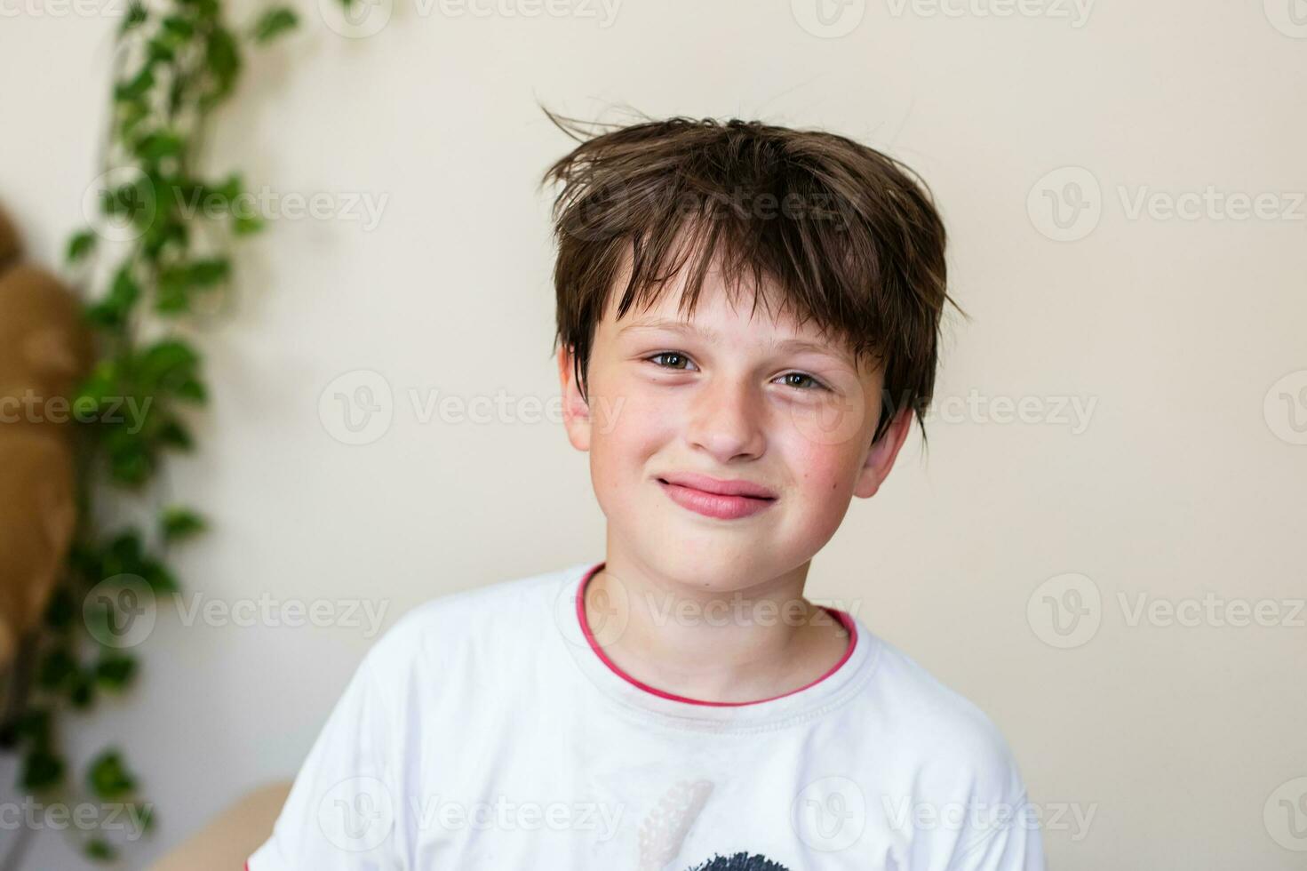 lindo adolescente Garoto às casa contra luz parede com tecelagem plantas. Garoto dentro branco camiseta e com desgrenhado cabelo sorri. Diversão pessoas. foto