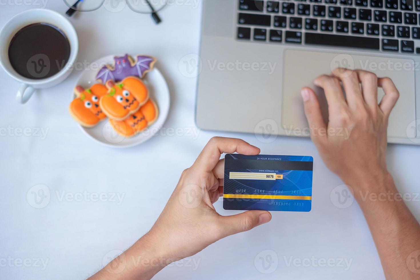 mão segurando o cartão de crédito para compras online no laptop durante comer café e biscoitos de halloween. feliz dia das bruxas, olá outubro, outono outono, conceito festivo, festa e feriado foto