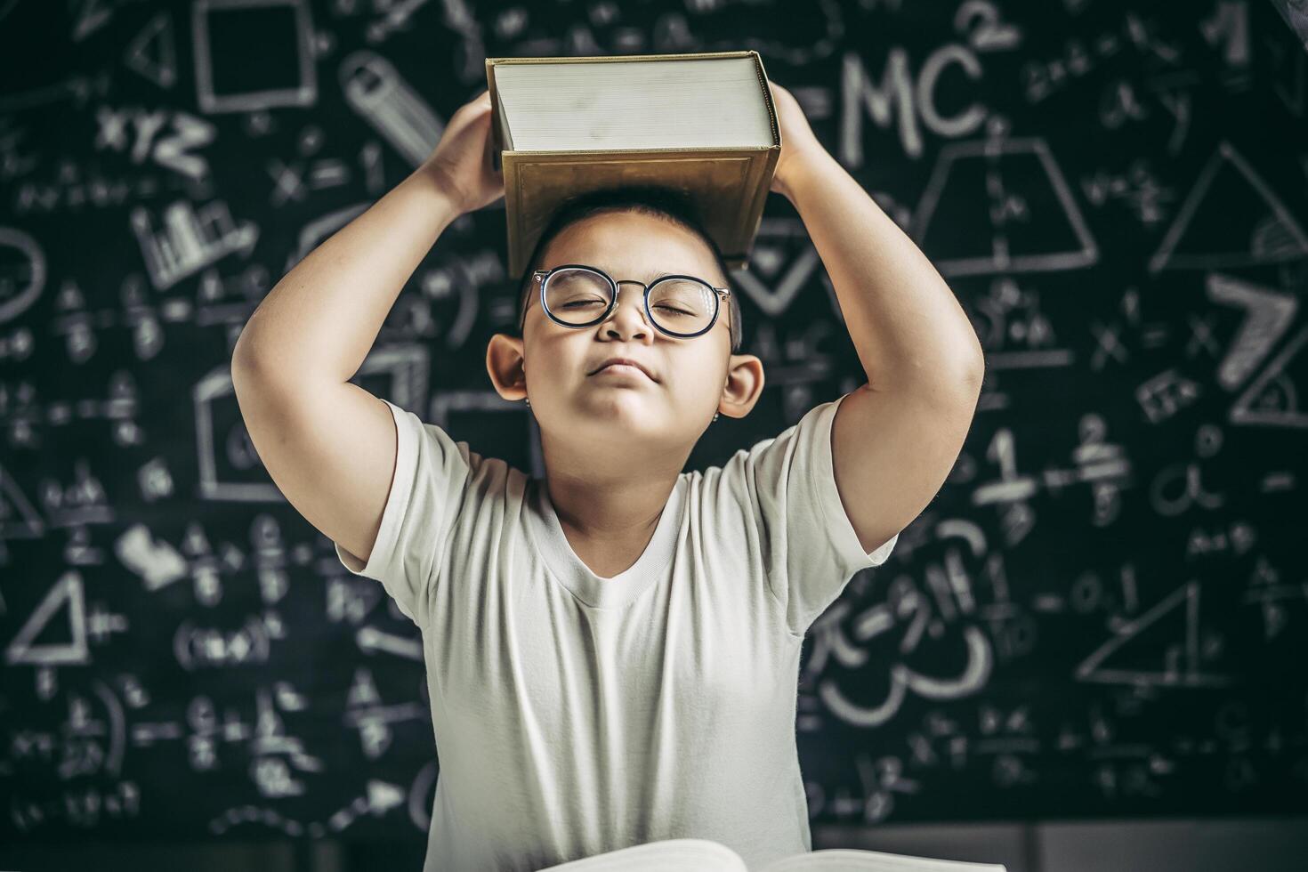 um menino de óculos estudava e colocava um livro na cabeça na sala de aula. foto
