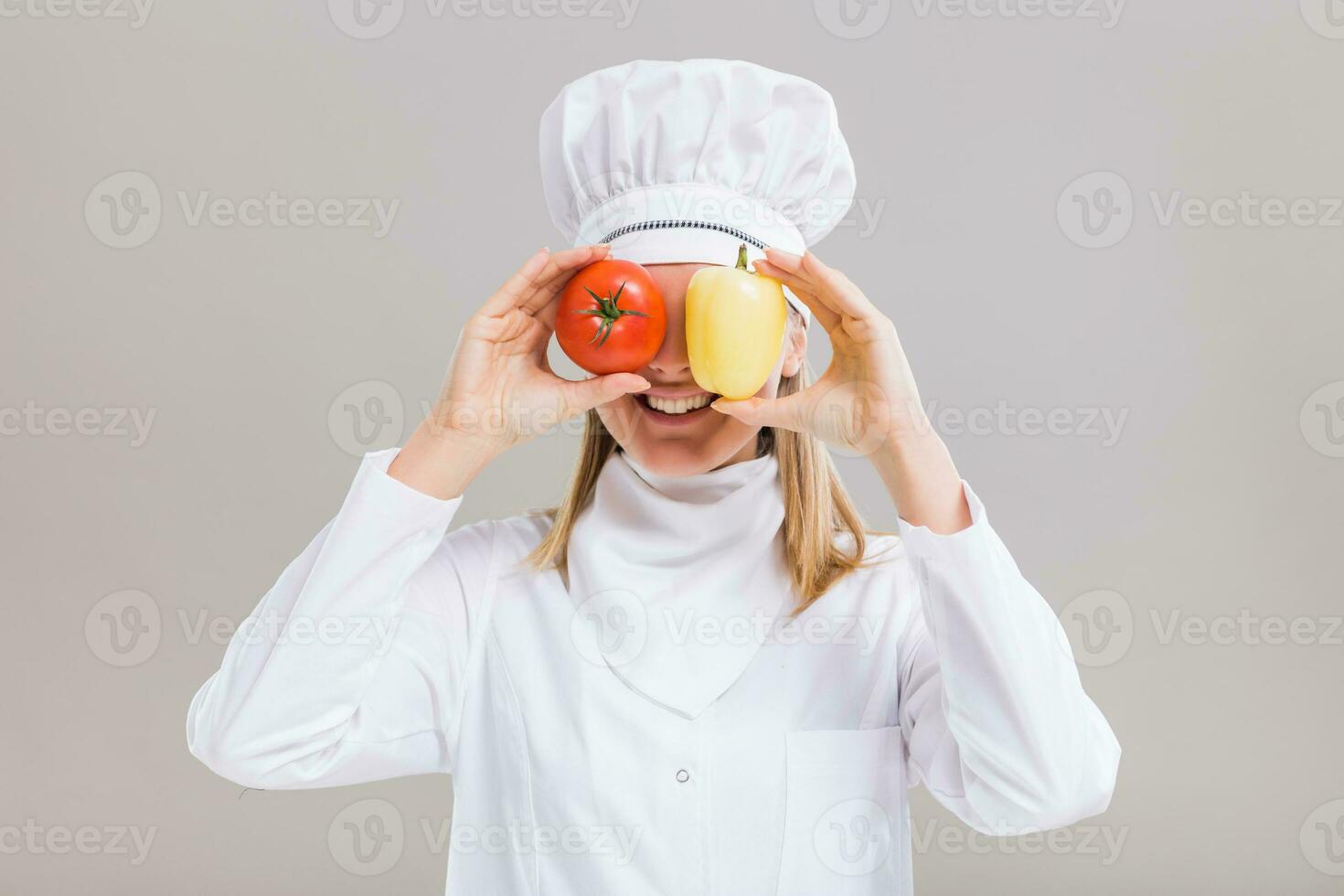 alegre fêmea chefe de cozinha é tendo Diversão com legumes em cinzento fundo. foto