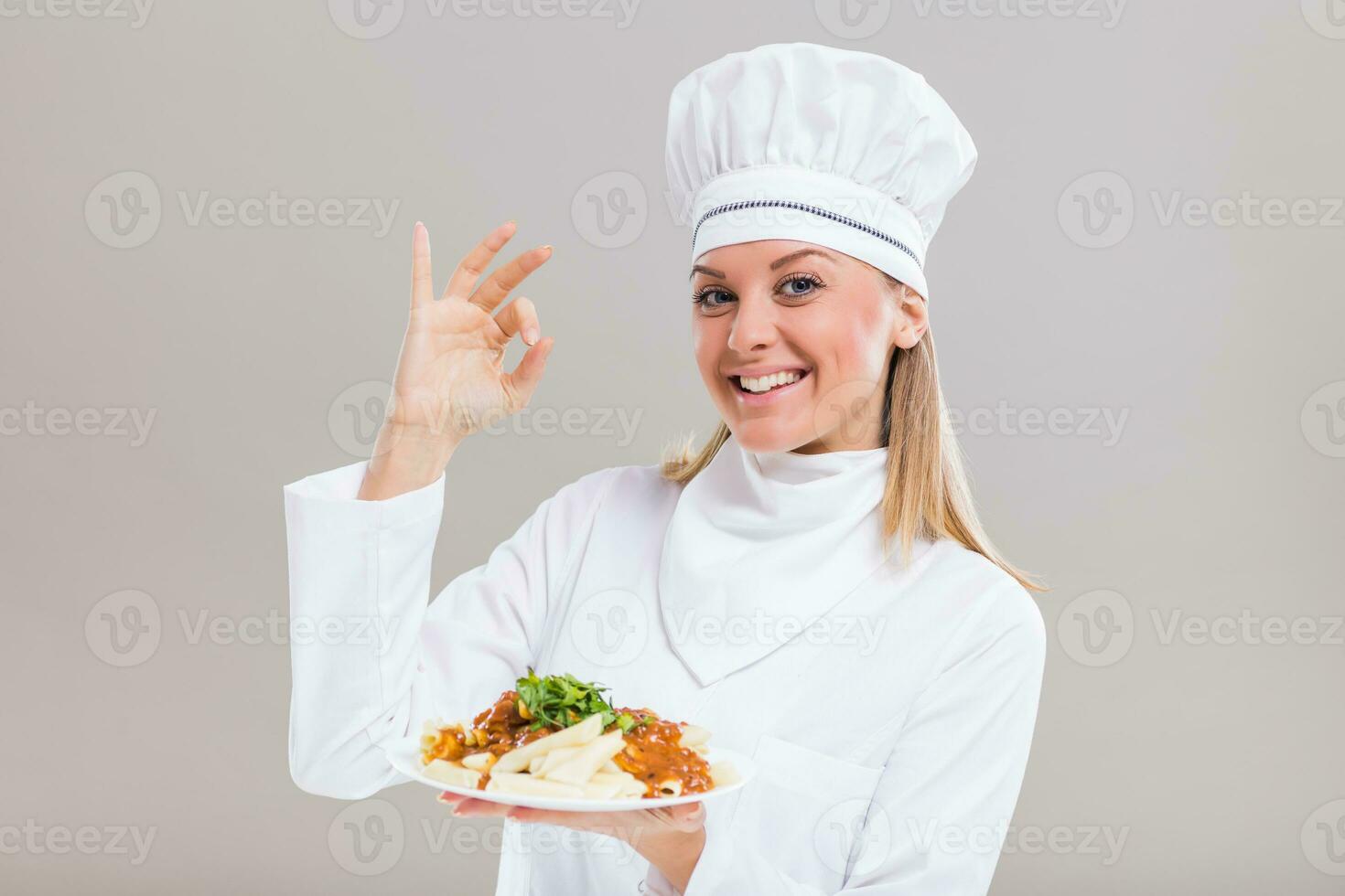 lindo fêmea chefe de cozinha é mostrando Está bem placa e preparado refeição em cinzento fundo. foto