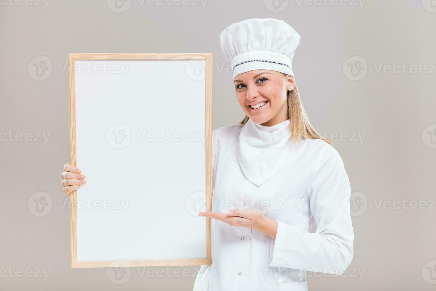retrato do lindo fêmea chefe de cozinha mostrando esvaziar quadro branco em cinzento fundo. foto