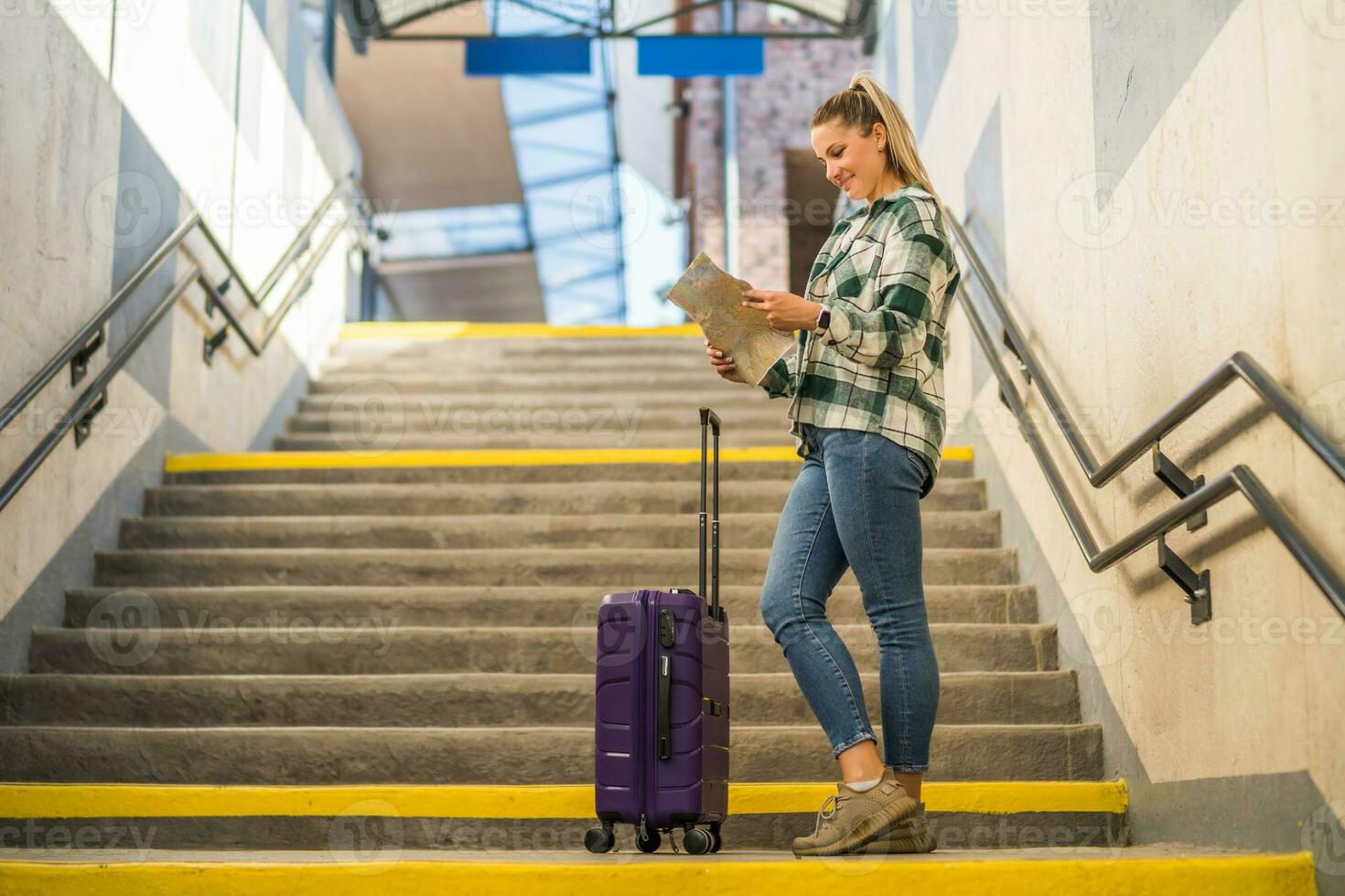 feliz mulher com mala de viagem olhando às mapa enquanto em pé em escadas do a trem estação foto