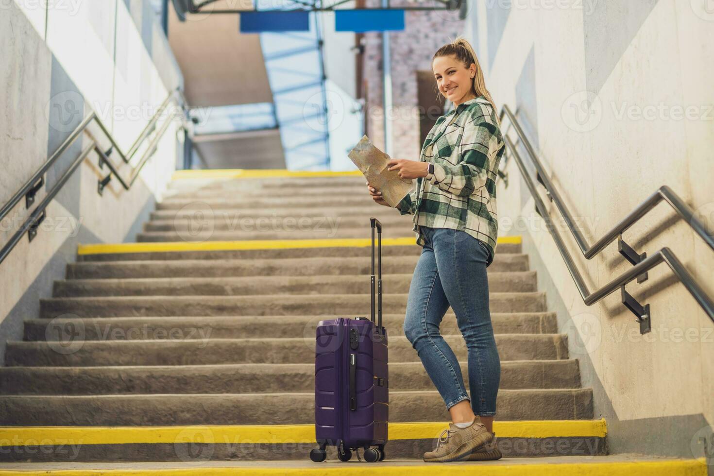 feliz mulher com mala de viagem olhando às mapa enquanto em pé em escadas do a trem estação foto