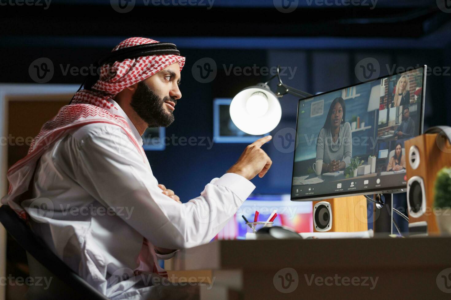 em computador, muçulmano cara é cuidadosamente ouvindo para dele colegas de trabalho durante grupo vídeo conversação. vista lateral foto mostra uma meio Oriental homem vestido tradicionalmente segurando a conectados reunião.