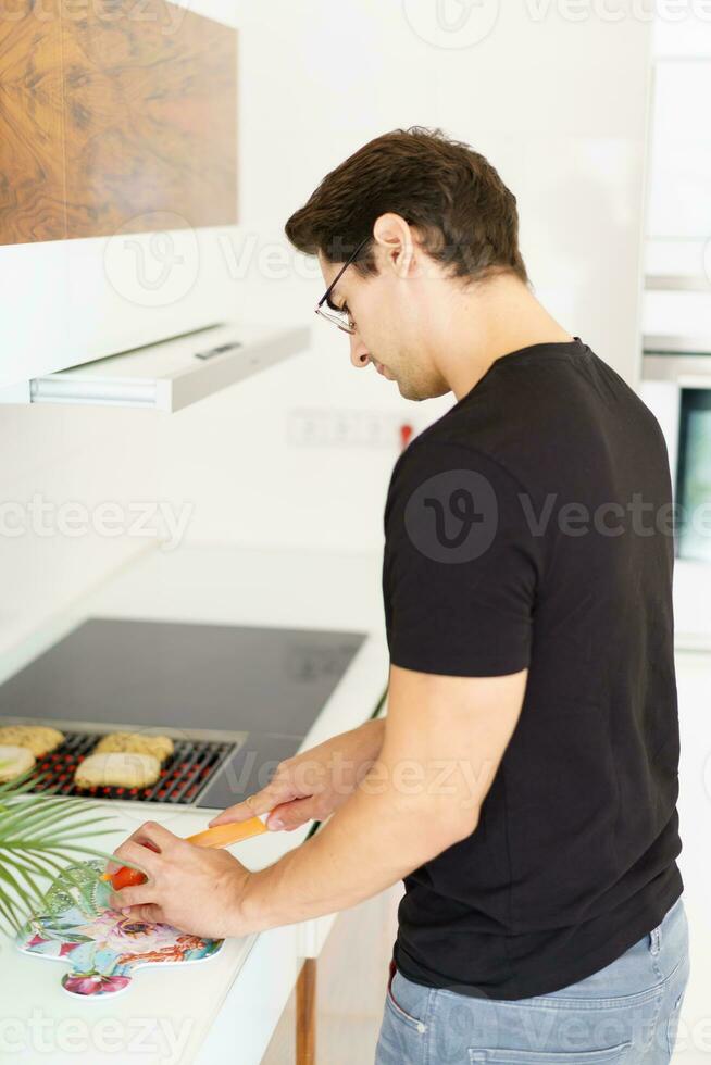 adulto homem em pé perto cozinhando alcance e corte tomate dentro cozinha foto