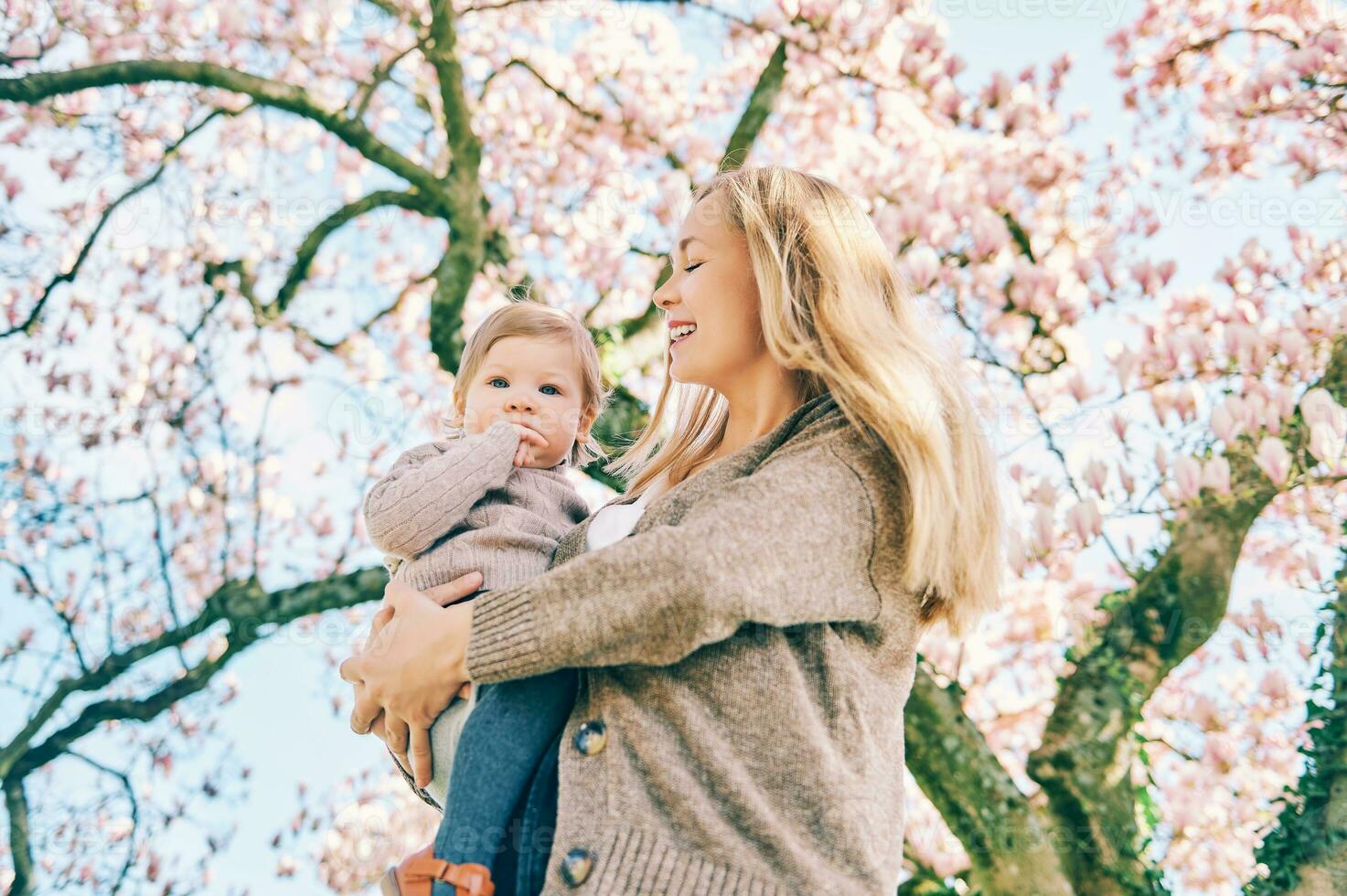 ao ar livre retrato do feliz jovem mãe com adorável bebê menina debaixo florescendo Primavera árvore, inferior Visão foto