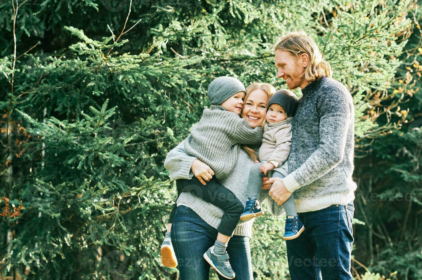 ao ar livre retrato do lindo feliz jovem família do 4 posando dentro pinho floresta, vestindo caloroso pulôveres, casal com criança pequena Garoto e bebê menina tendo Boa Tempo às natureza, frio clima foto