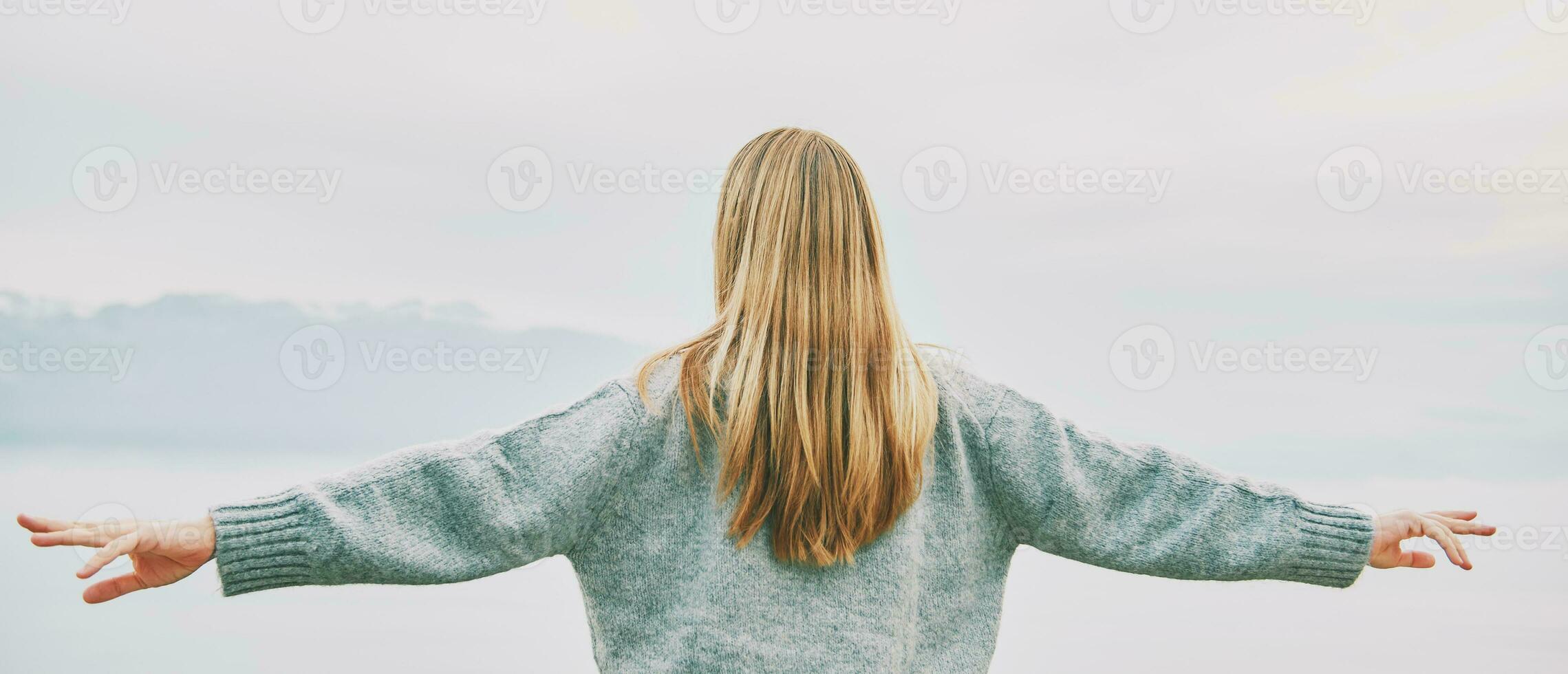 panorâmico bandeira fundo costas Visão do jovem loiro mulher com braços Largo aberto apreciar panorama do nublado montanhas foto