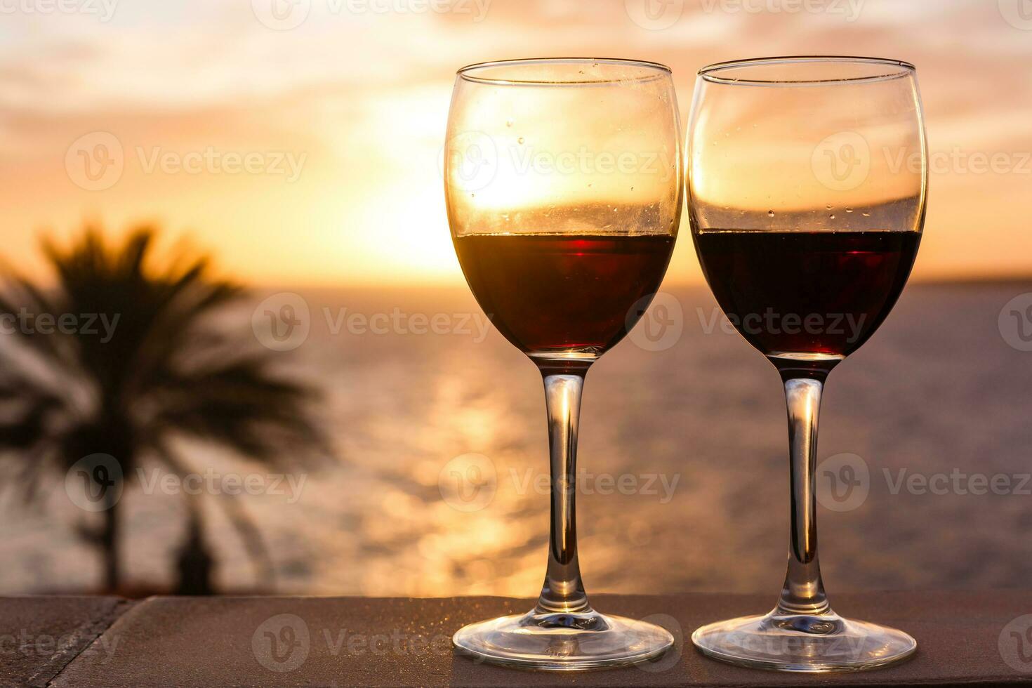 dois óculos do vermelho vinho sentado em uma borda sobre olhando a praia, oceano e uma lindo pôr do sol. foto