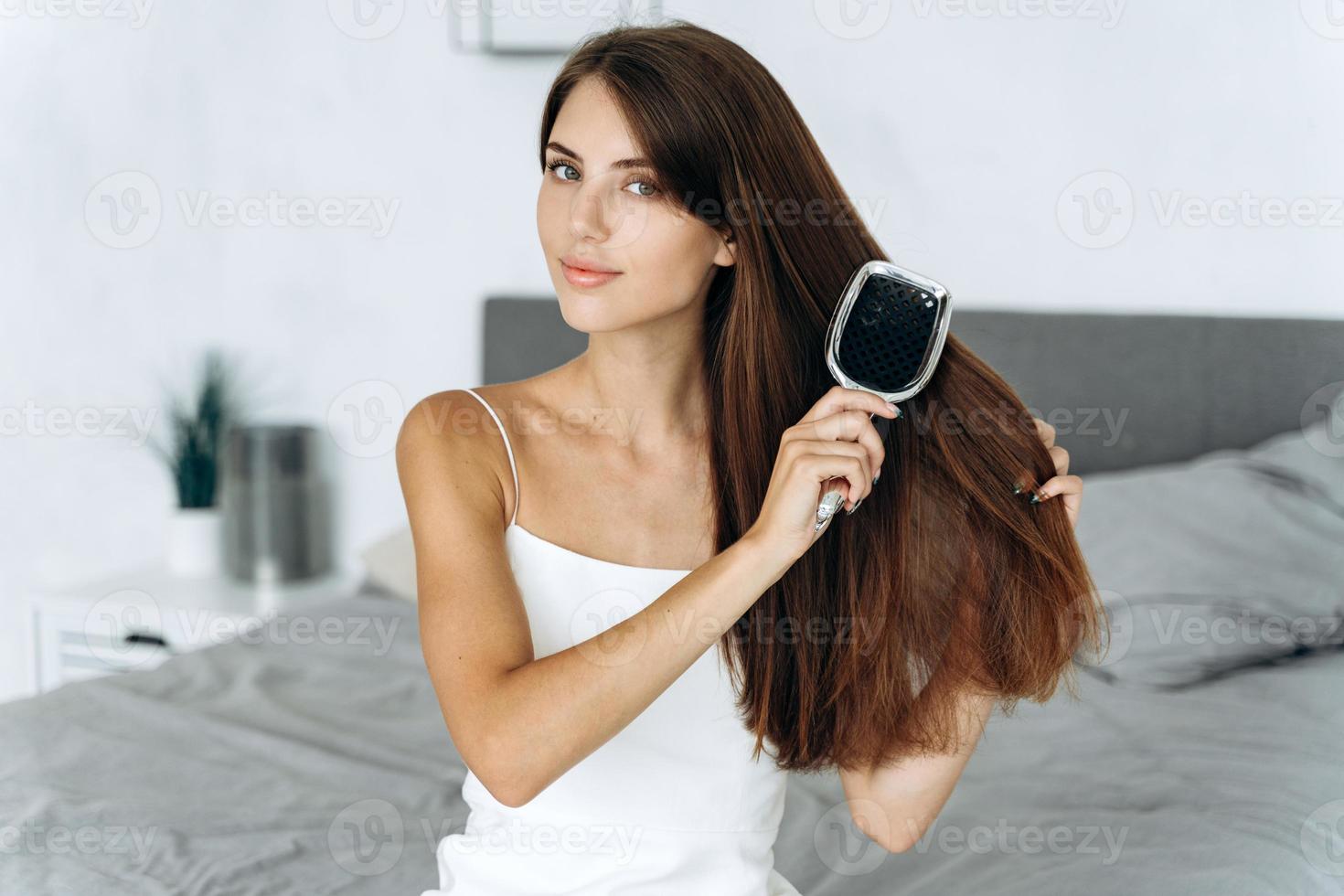 mulher morena saudável, escovando o cabelo com o pente antes da caminhada, enquanto está sentado no quarto e olhando para a câmera. conceito de procedimentos de autocuidado foto