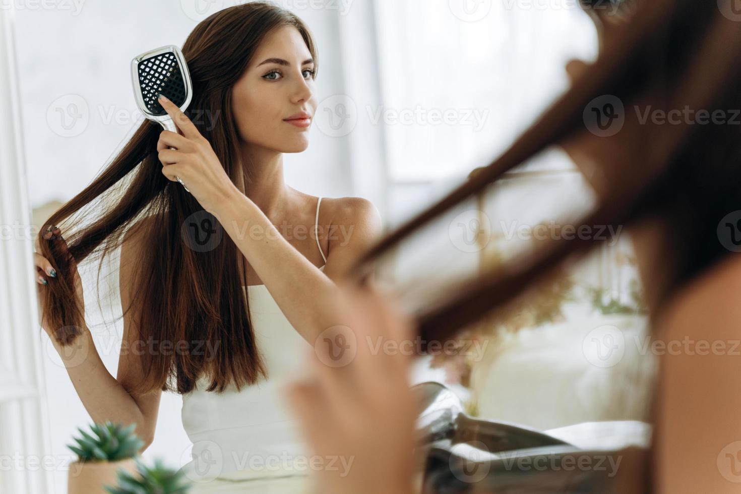 cintura para cima vista do retrato da mulher bonita feliz em roupa doméstica, penteando o cabelo na frente do espelho, segurando uma escova grande. foto