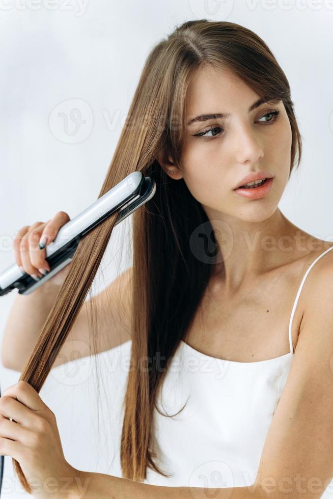 visão vertical de uma jovem bonita e alegre caucasiana passando a ferro o cabelo dela em casa enquanto desvia o olhar e pensa em algo foto