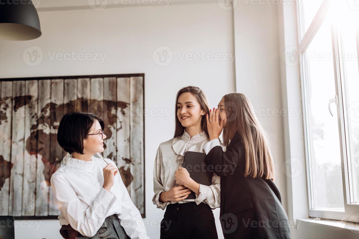 conceito de trabalho, questões polêmicas. três garotas confiantes conversando no escritório, uma sussurra no ouvido da outra foto
